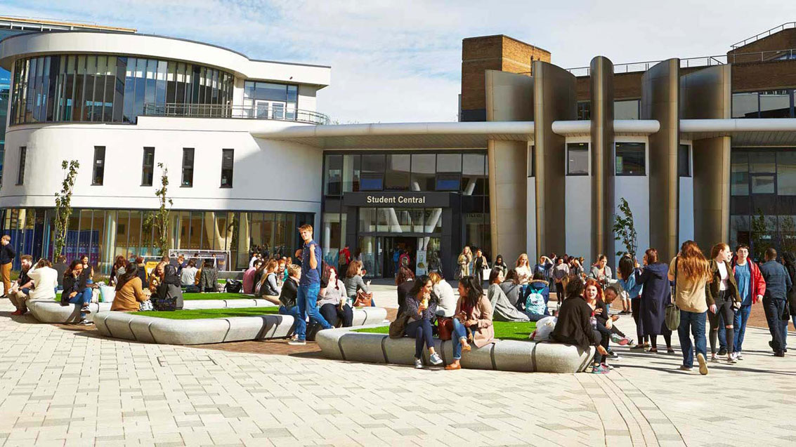 A sunny day in University square. Students are sat out on the blocks and the image point of view is looking at Student Central.